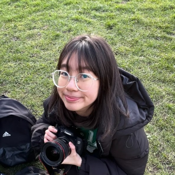 Kher Shin a Malaysian student wears round glasses and smiles up at the camera, whilst also holding a camera.