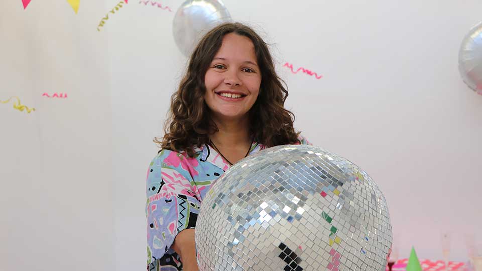 Young white female with long dark brown hair smiling at the camera and holding a large glitterball.