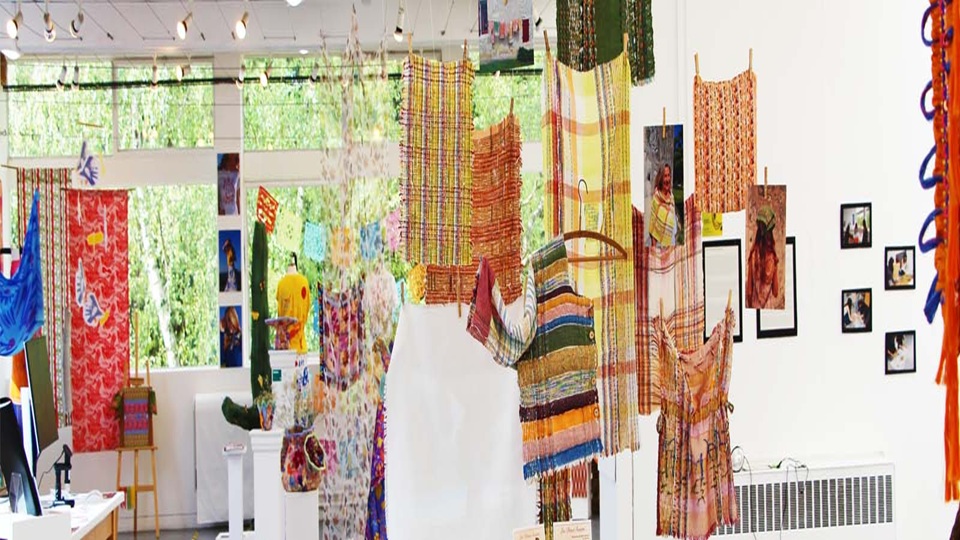 Textiles exhiibition with various woven fabrics in bright colours and patterns hanging from the ceiling with a backdrop of bright windows along the back wall