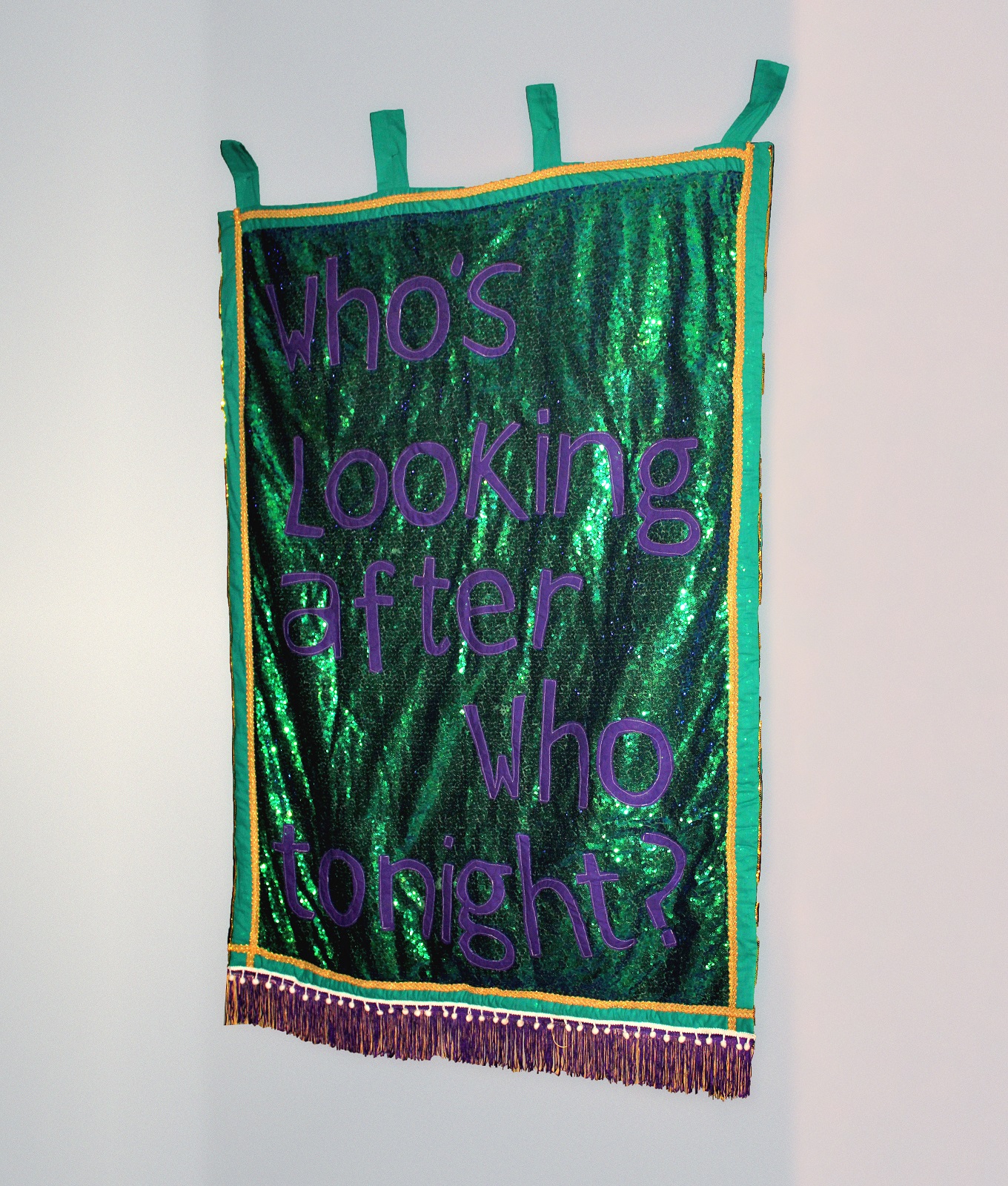 A green sequined banner on a white wall saying Whos Looking After Who Tonight