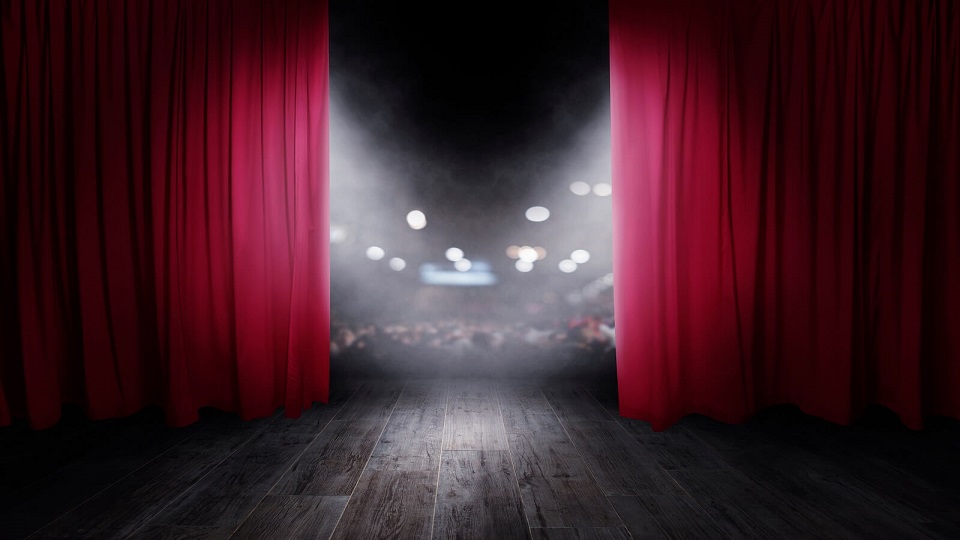 A theatre stage dimly lit looking towards the audience from behind half opened red stage curtains