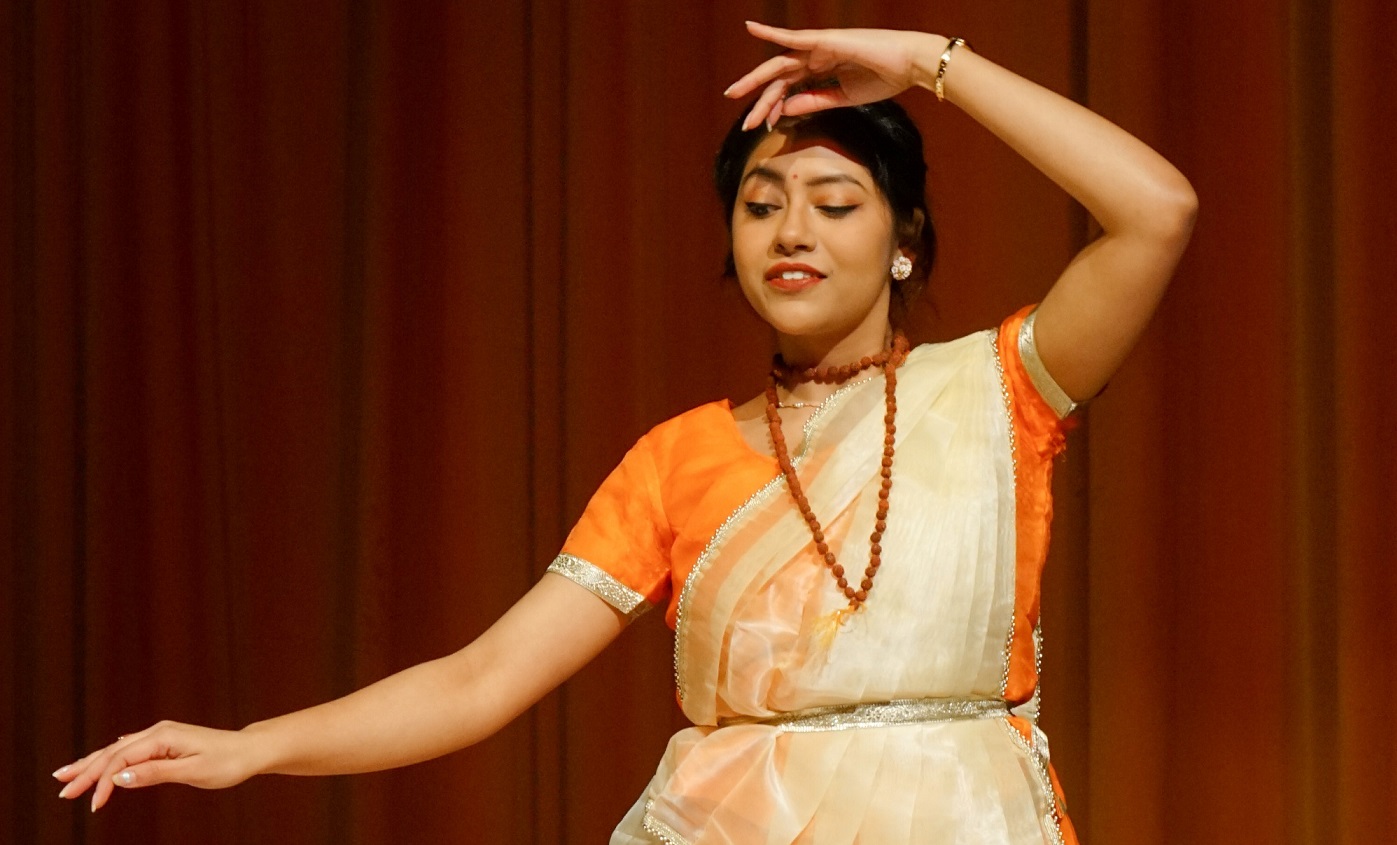 A young woman in her early 20s of indian heritage dances in a traditional saree in front of an orange curtain