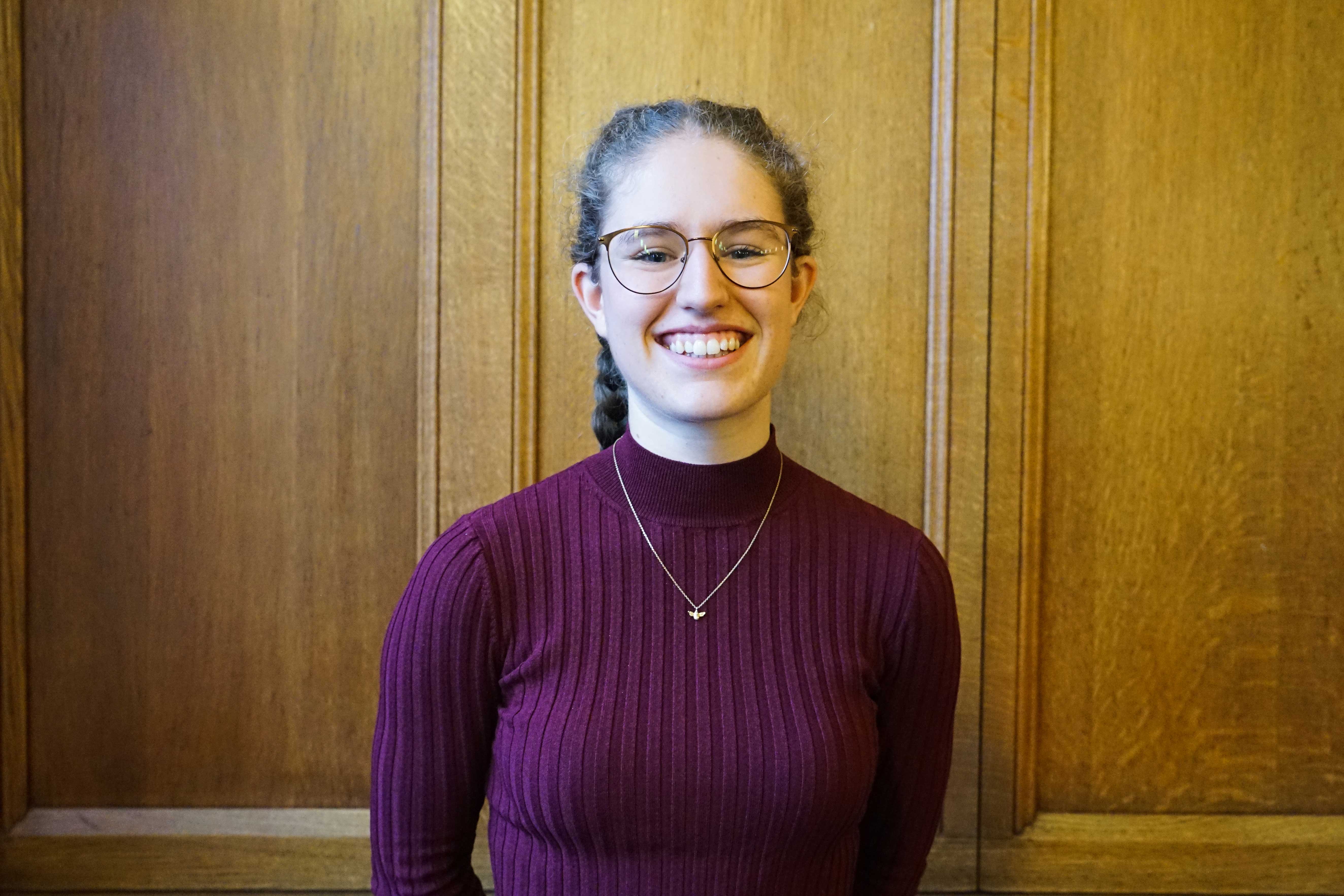 White female student in late teens/early twenties, smiling at the camera. She has a dark red long sleeve top on with a fine chain gold necklace. Her long brown hair is tied back from her face and she wears glasses.