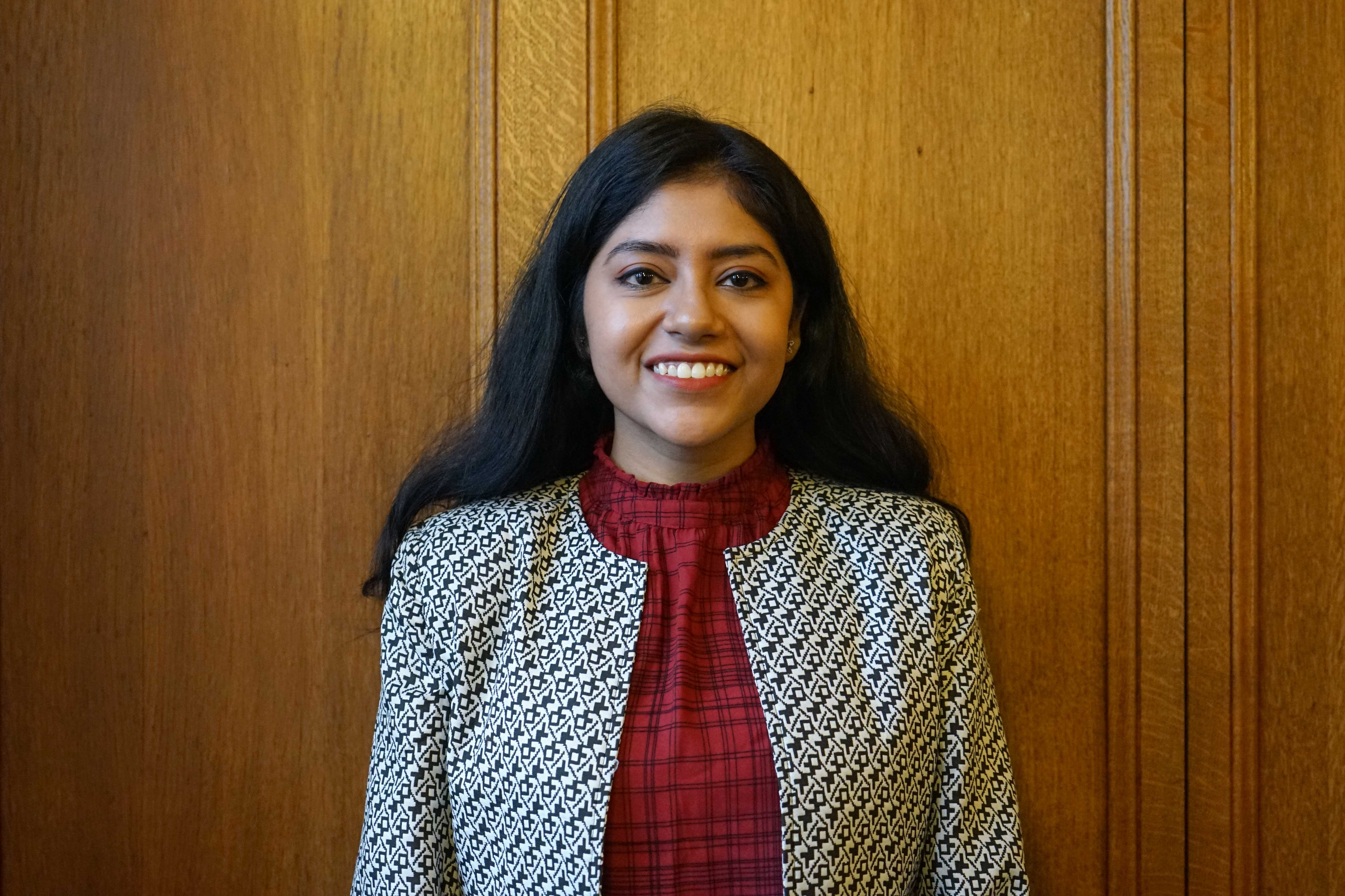 Female mature student in her twenties with light brown skin smiling at the camera. She is wearing a red top under a black and white patterned jacket and has long straight black hair.