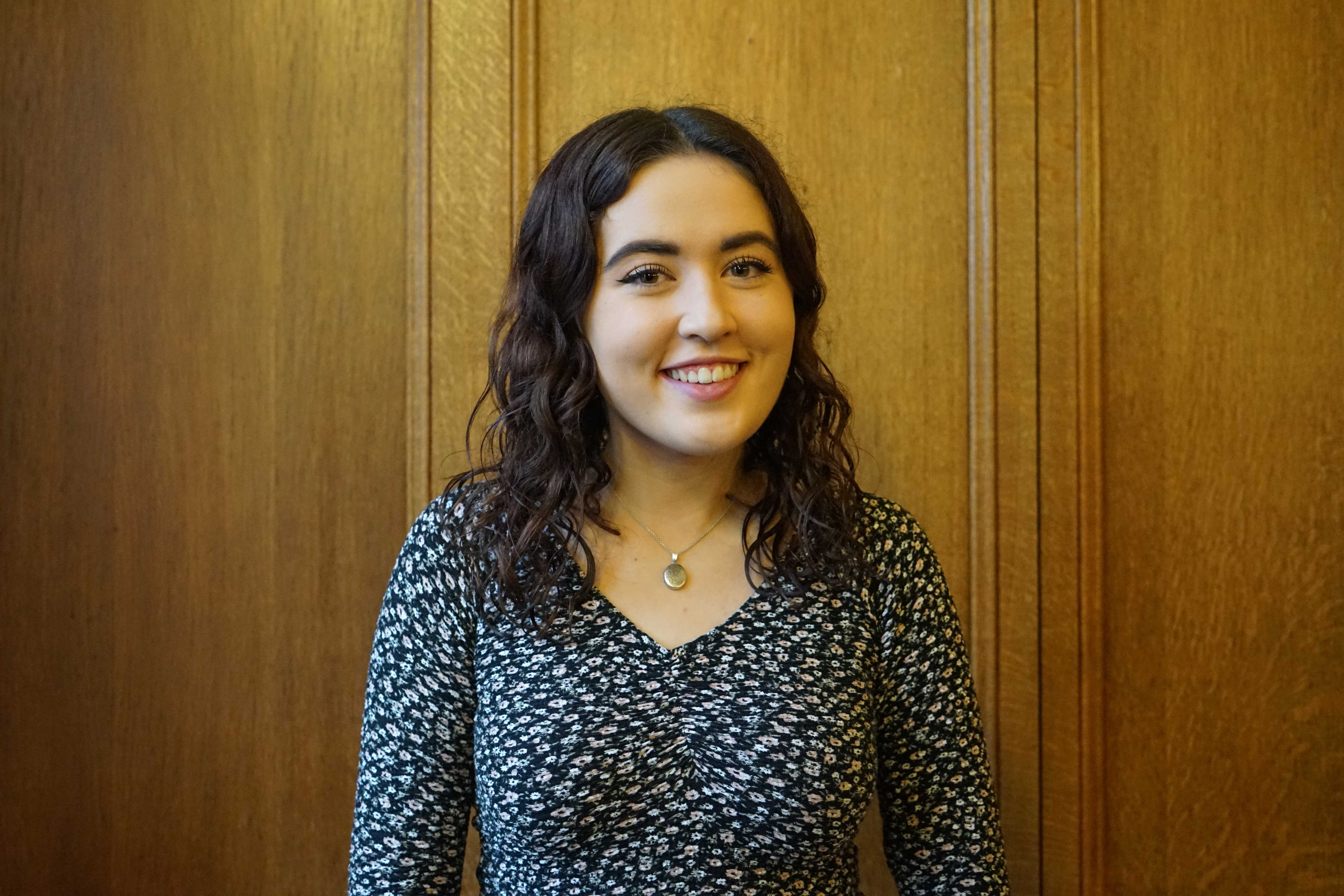 White female student in her late teens/early twenties smiling at the camera. She has long curly dark brown hair and is wearing a black and white top with a small floral print and a necklace.