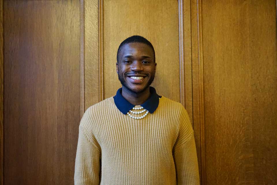 Black male student in his late teens/twenties smiling at the camera. He has short cropped black hair and is wearing a pale mustard colour jumper with a black collar visible and a three layered pearl necklace.