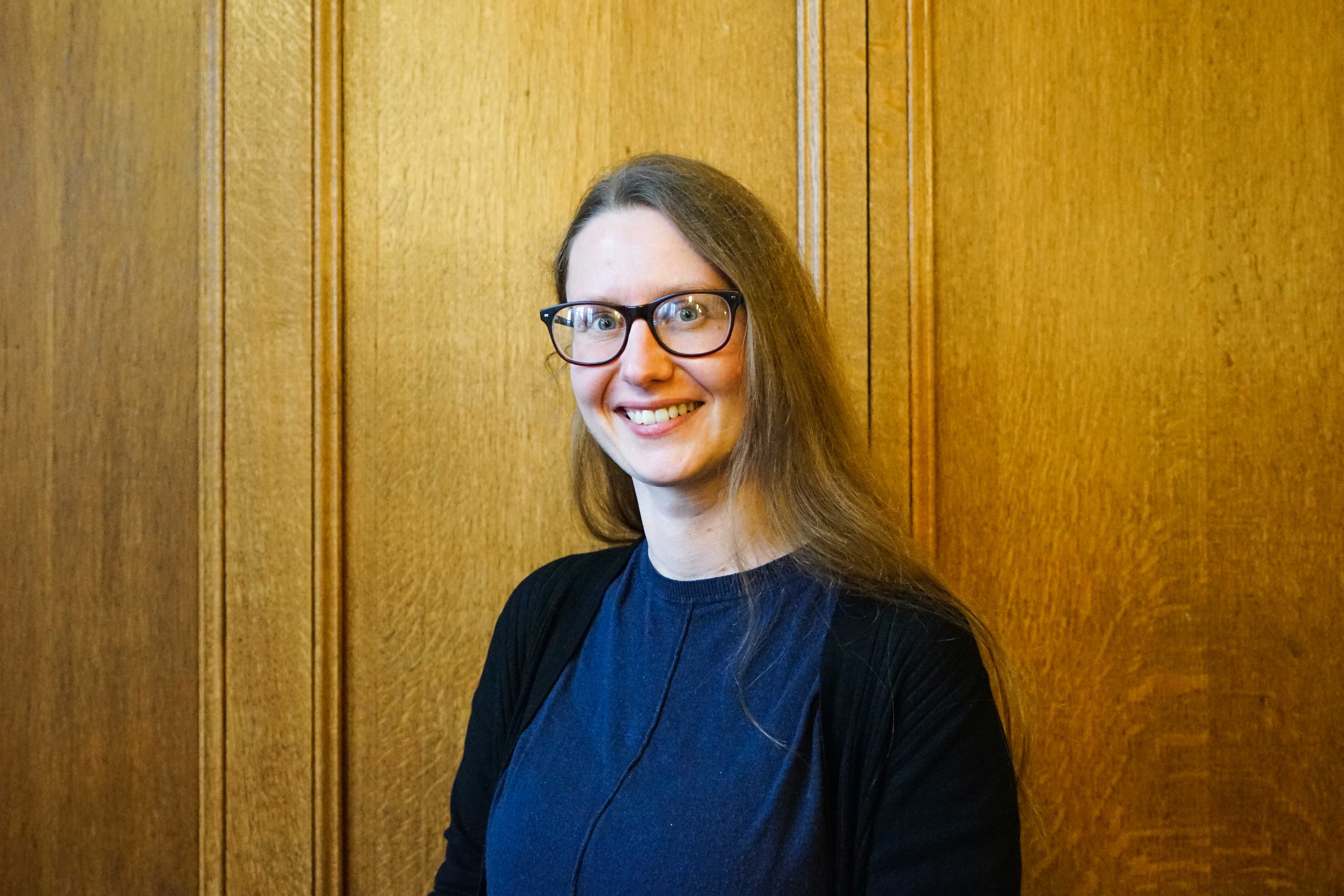 A white female student in her early forties, wearing a dark blue top and black cardigan. She is smiling and has long straight brown hair and is wearing glasses.