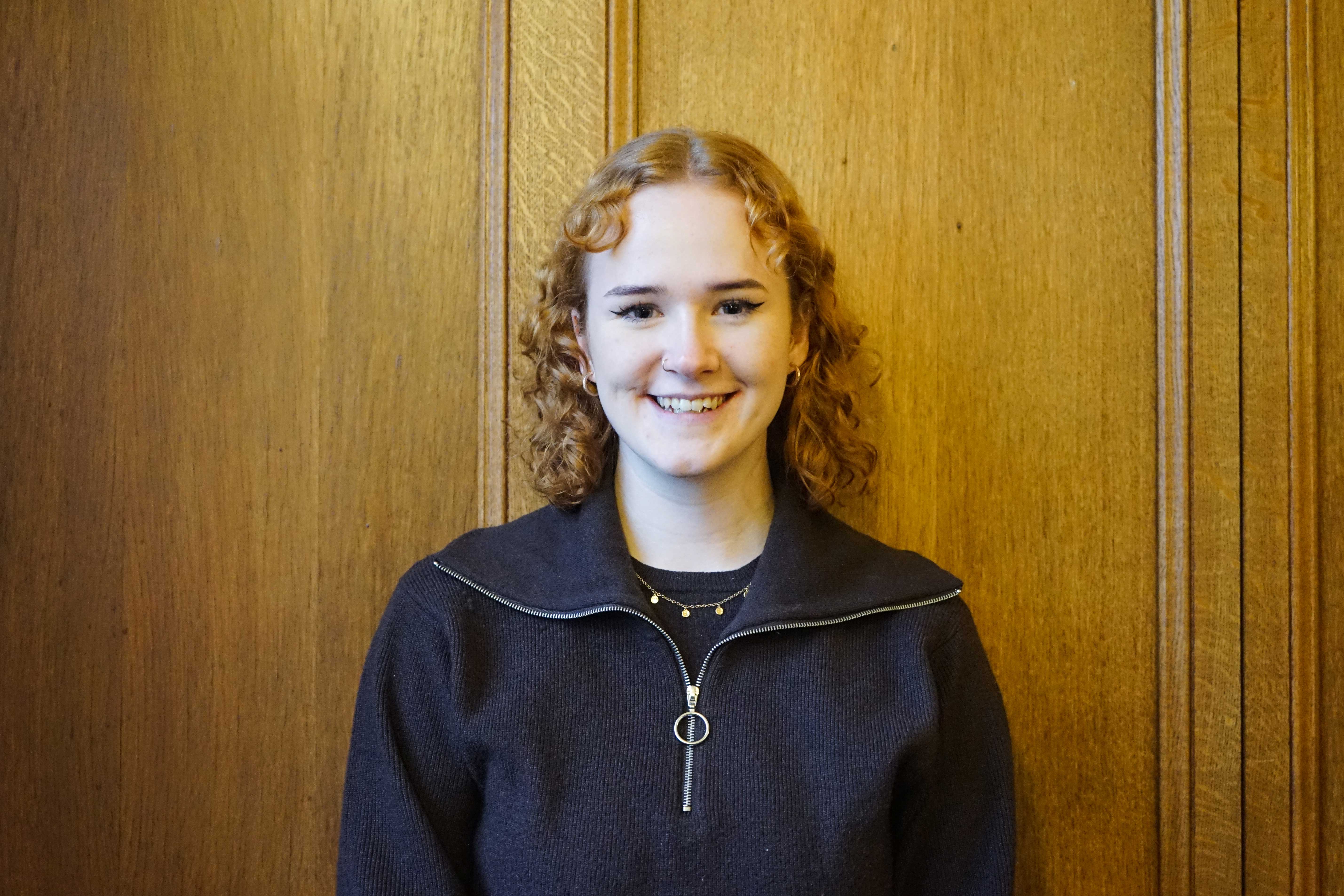 A white female student in her early twenties, wearing a dark top with a zip collar. She has medium length red curly hair and is smiling at the camera.