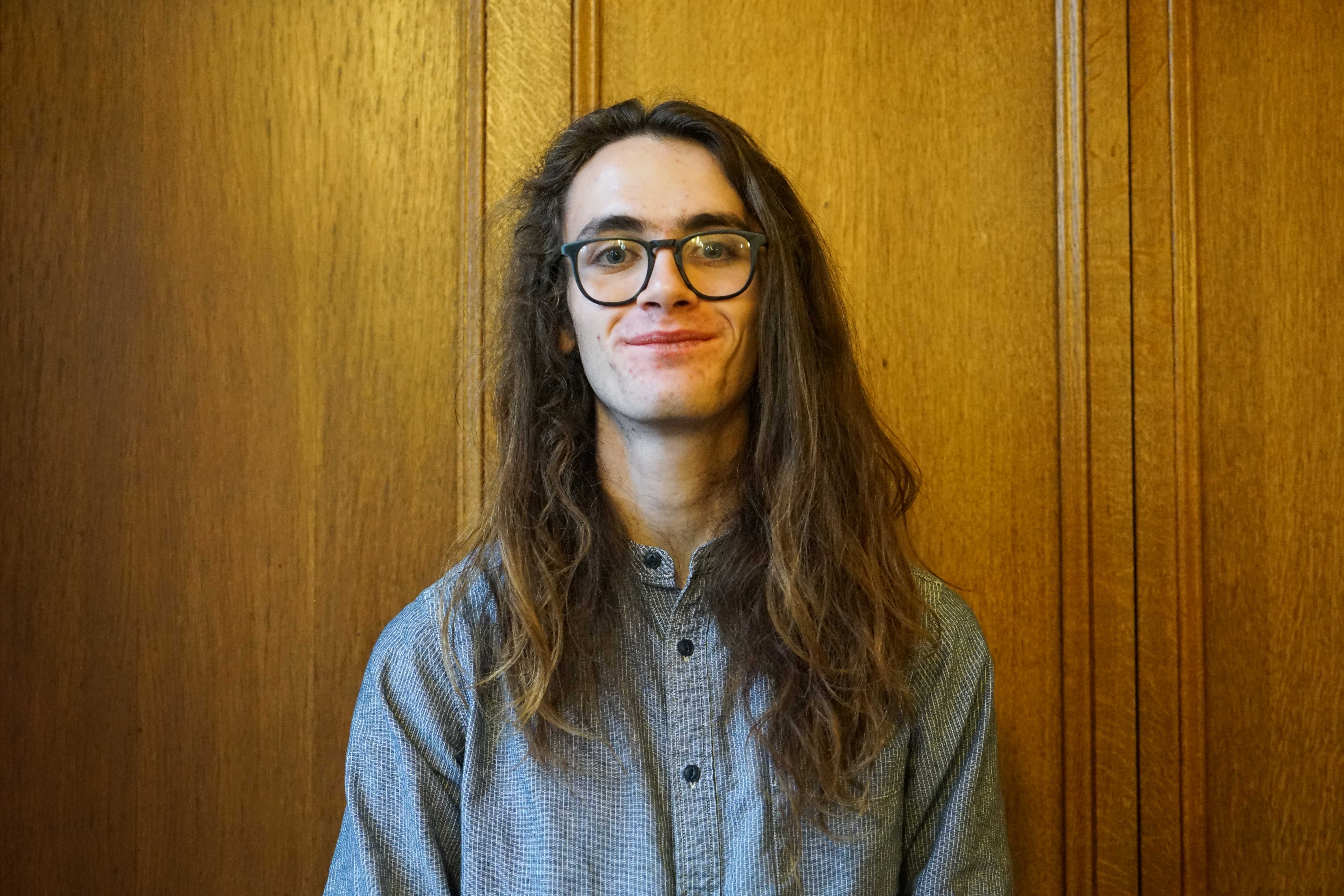 A white male student in his twenties with very long dark brown hair. He is wearing a grey shirt and glasses.