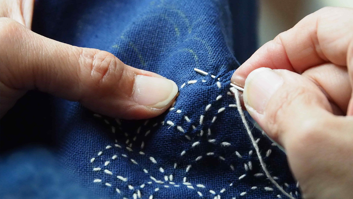 Close up of a hand sewing white thread on blue fabric.