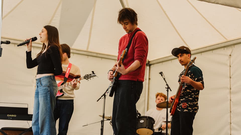 A student band with three male guitarists, a drummer behind them and a female singer at the front. They are playing in a white marquee.