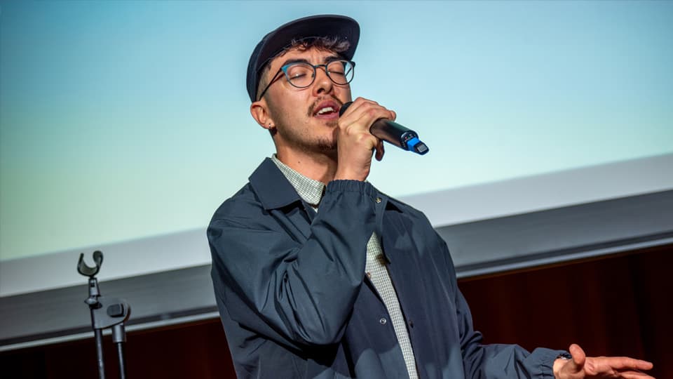A man wearing a blue jacket, cap and glasses, singing into a microphone with his eyes closed. There is a blue background behind him.
