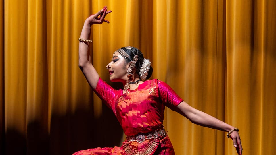A woman dancing in traditional Bharatanatyam dress in red and gold, with one arm raised above her head. There is a yellow curtain behind her.