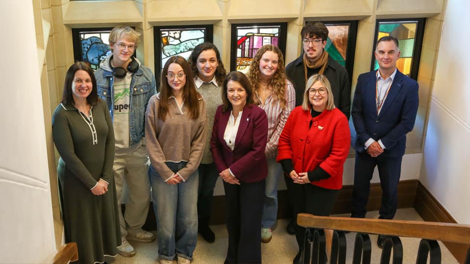 A group of students and staff stood smiling in front of a stained glass window.