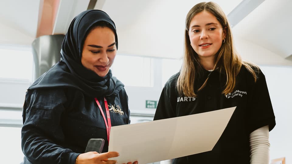 A woman wearing a black hijab, looking down at a sheet of white paper and smiling. A woman with long brown hair is stood next to her, also smiling.