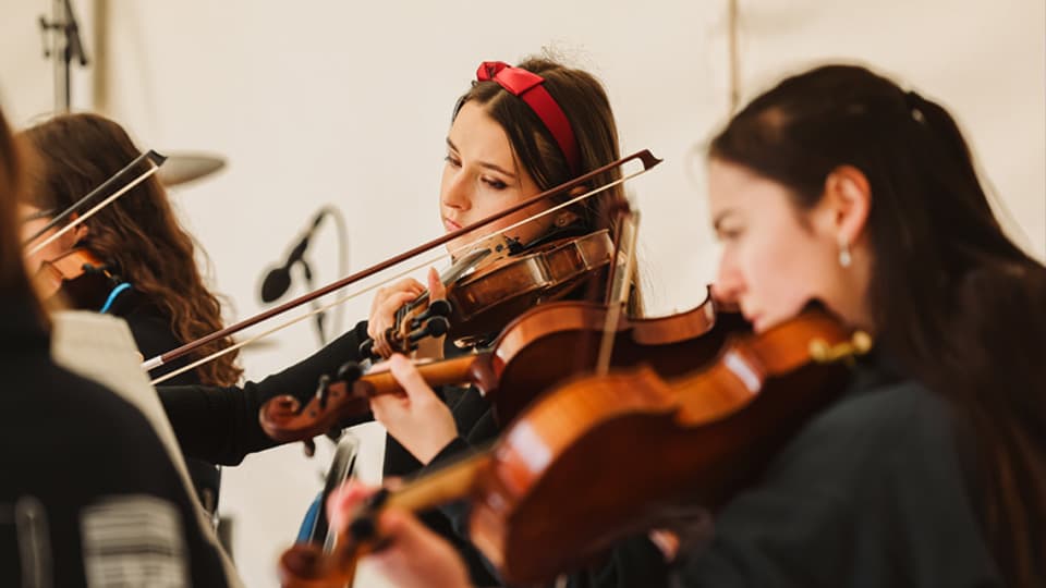 A woman with long brown hair and a red bow headband, playing a violin and reading from a black music stand.