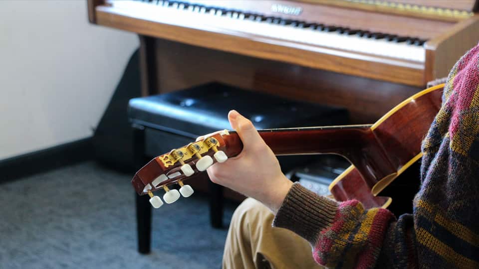 Close up of a man wearing a red patterned jumper, playing a guitar in front of a brown piano.