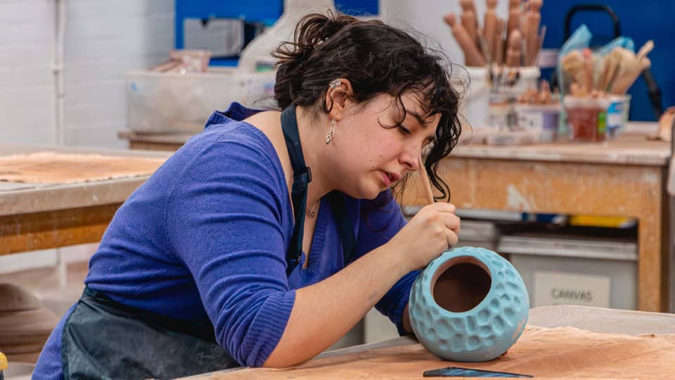 A woman with brown curly hair, wearing a blue top and a black apron. She is looking down and painting a small blue ceramic vase.
