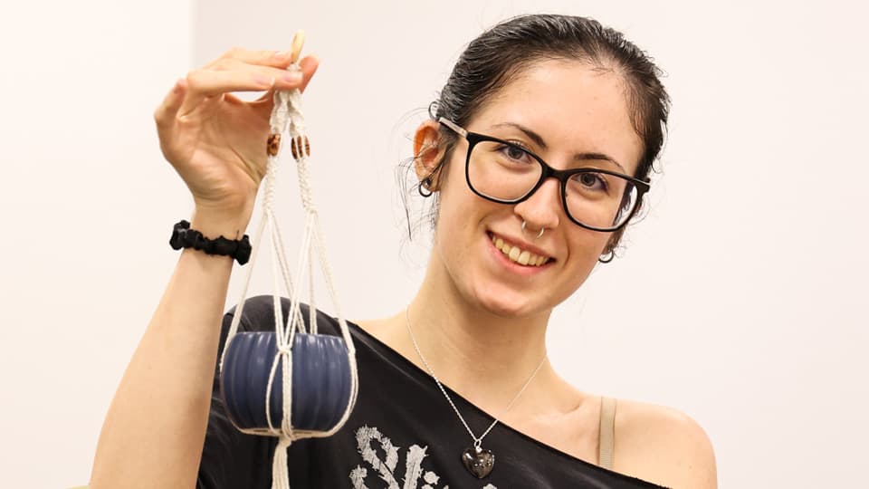 A woman with brown hair tied back and black glasses, smiling and holding up a small blue plant pot, suspended in a macrame plant hanger.