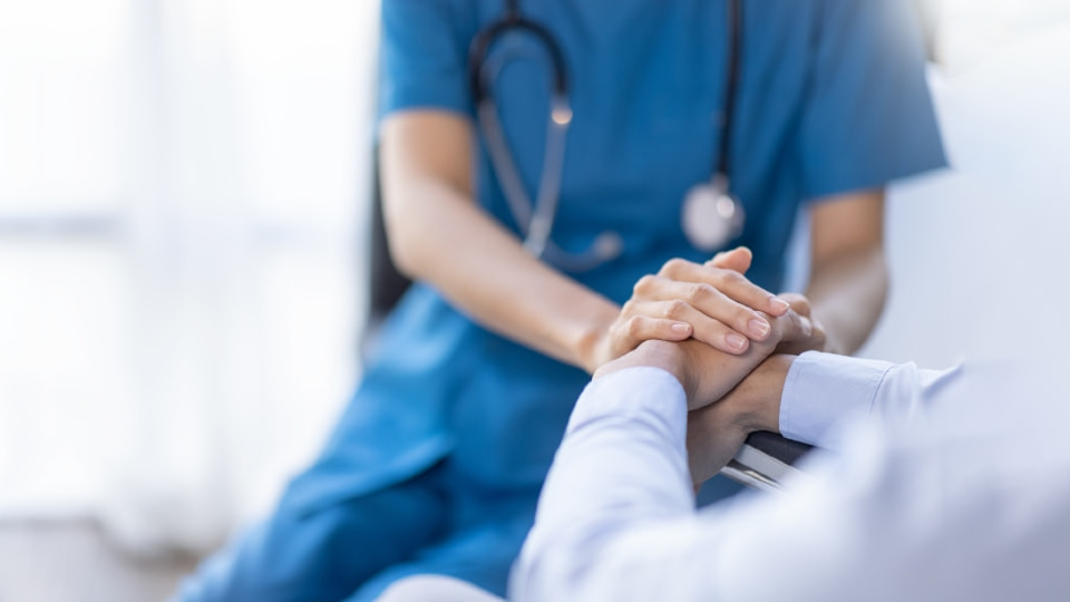 A nurse in a blue uniform with a stethoscope around their next holding a patient's hand