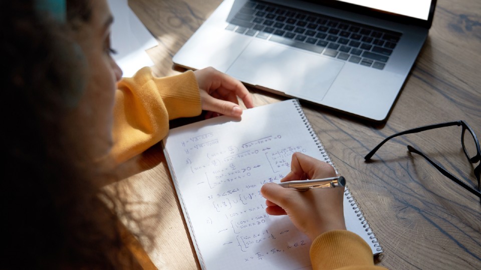 Picture of a teenage girl in a yellow jumper writing down maths equations in front of a laptop