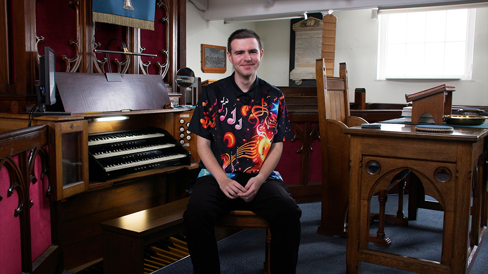 Thomas Corden seated, wearing a shirt with music notes on it. Behind him is an organ.