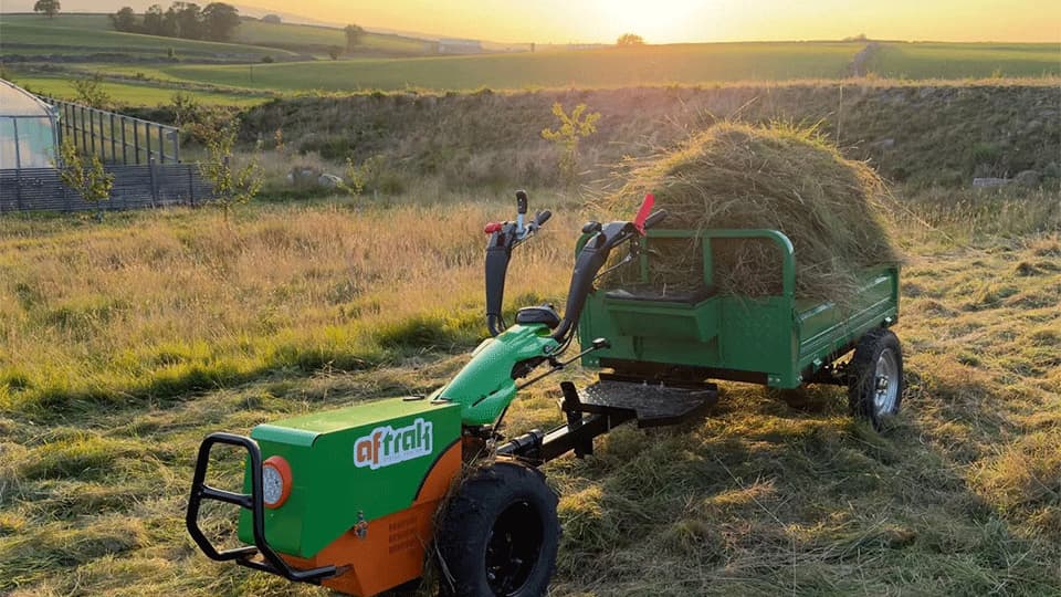 Aftrak tractor and trailer loaded high with cut grass, standing in a mown meadow at sunset with the Dales behind