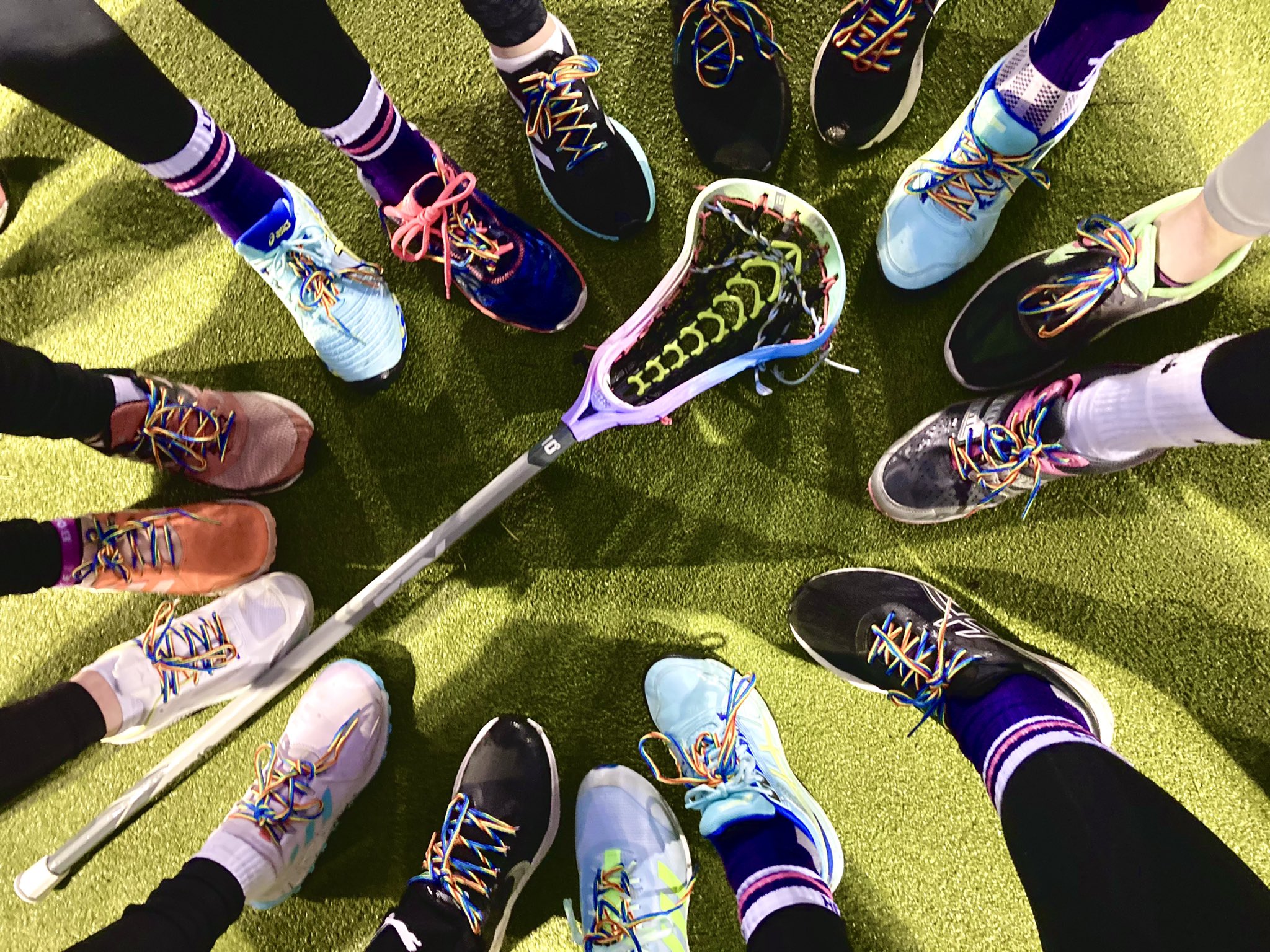 sixteen shoes laced up with rainbow laces and a lacrosse stick in the centre