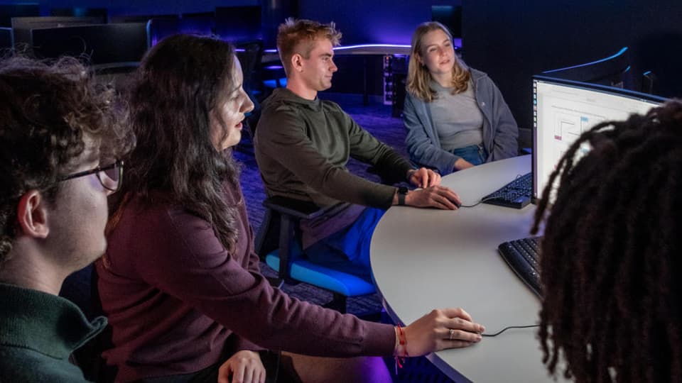 A group of people sat in a dark room working at a bank of computers.