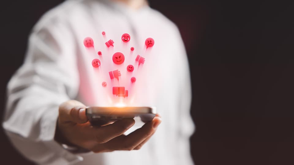 A person in a white sweatshirt holding a mobile phone with red emoji's emerging from the screen.