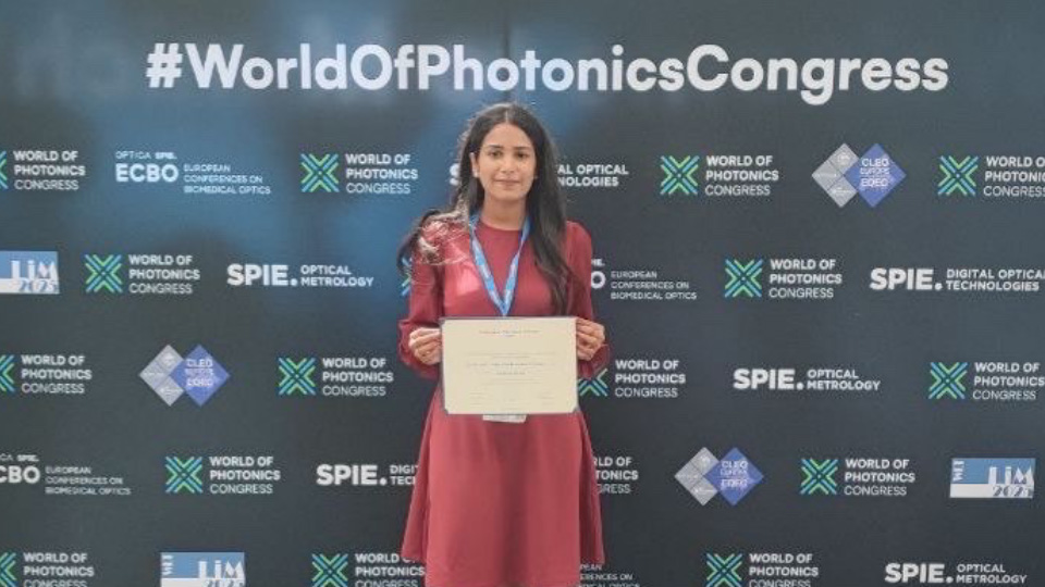 A woman stands holding a certificate in front of a backdrop with logos and the text World Of Photonics Congress.