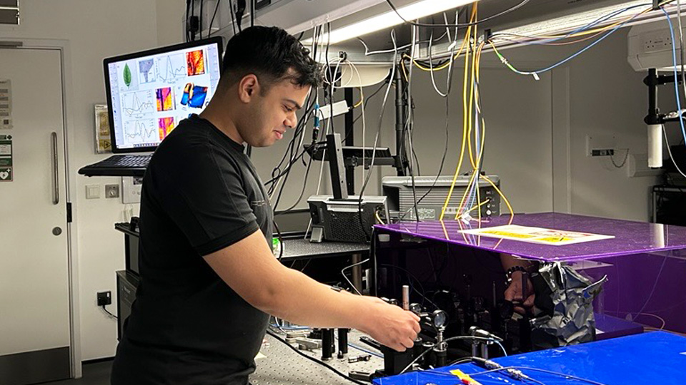 A man adjusts equipment in a laboratory filled with electronic devices, cables, and a computer displaying scientific diagrams.