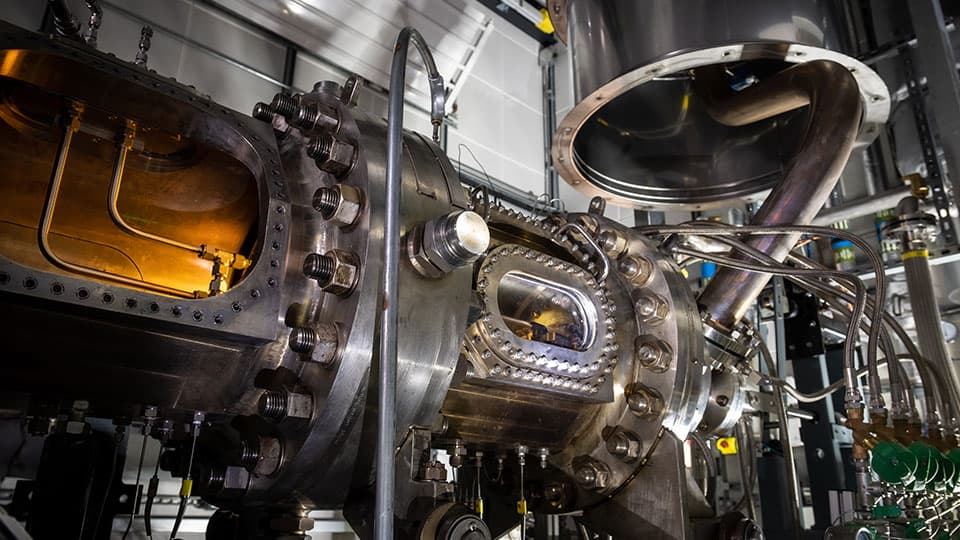 An industrial, heavy-duty metallic machine assembly features complex piping, bolted flanges, and small viewing windows revealing a lit amber-colored interior.