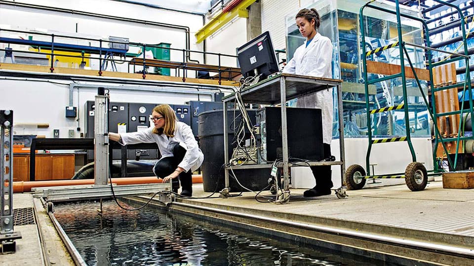 Two scientists work with specialized equipment and a computer station next to a large water flume tank in a hydraulics laboratory.