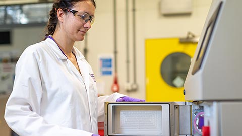 A female researcher in a white lab coat, eye protectors and purple gloves, opening the door of a fume cupboard