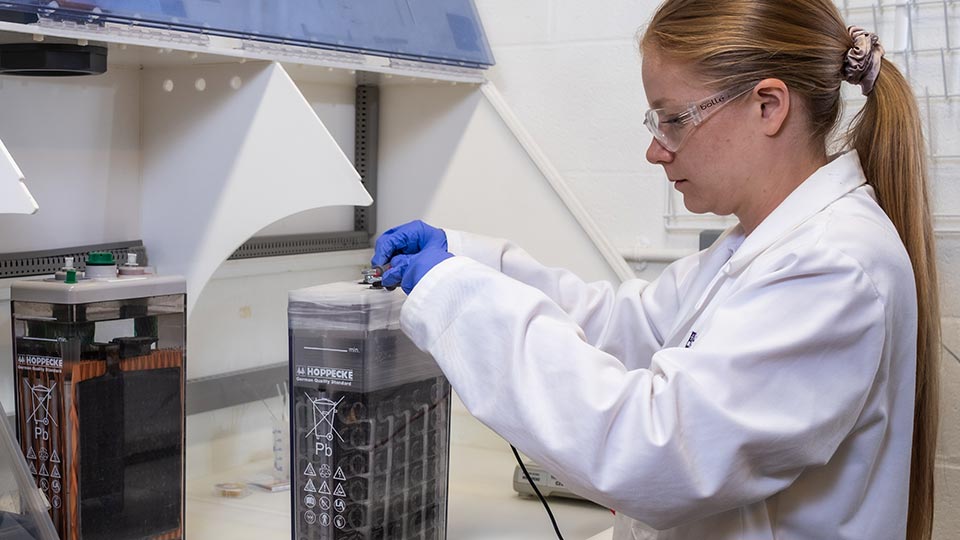 A female researcher in a white coat, wearing protective eyewear and blue gloves connecting wires to a battolyser