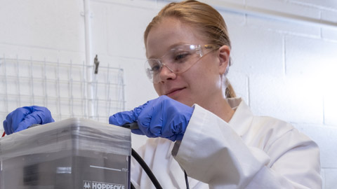 A female researcher with safety glasses, white lab coat and blue plastic gloves connecting wires to a battery