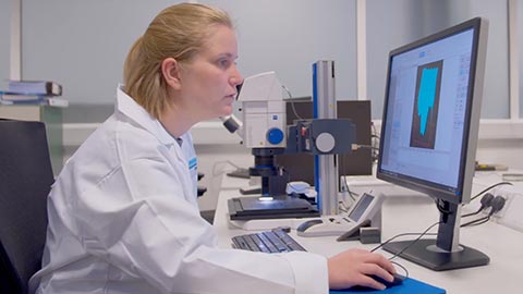 A female researcher in a while lab coat sitting at a desk with a monitor on it, with one hand operating a mouse
