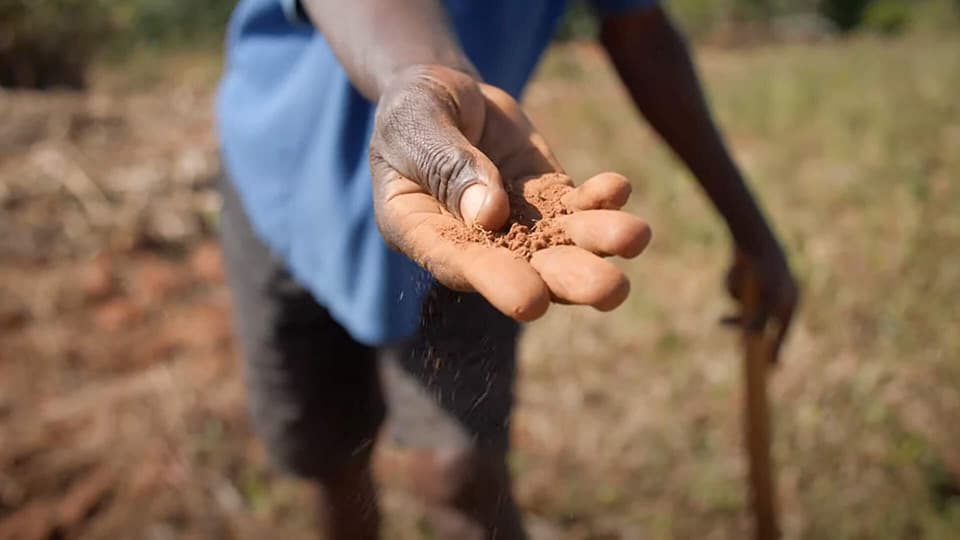 A close-up of a person's hand holding some soil, with a blurred field and a wooden tool visible in the background.