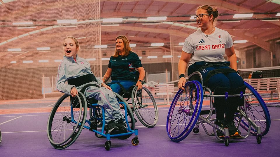Three people in wheelchairs positioned on a purple indoor court, actively participating in a sports activity together.