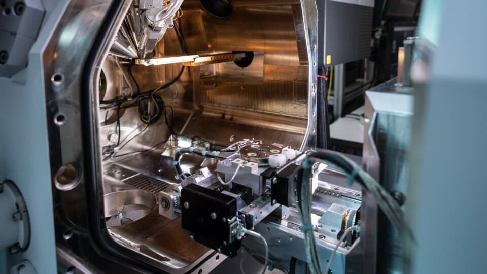 Close-up view of scientific equipment inside a laboratory, featuring metal components and wiring within a large stainless steel chamber.