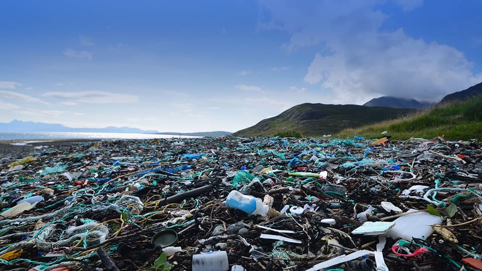 A rocky beach covered in plastic waste, fishing nets, and debris, with mountains and a blue sky with clouds in the background on a sunny day.