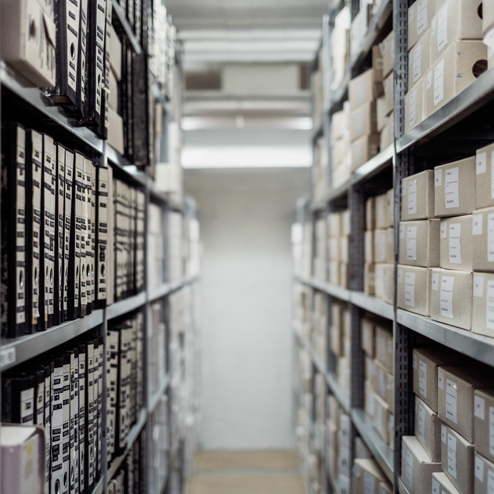 Photograph looking down the length of the walk space between two stacks in a traditional archive - full of books and files