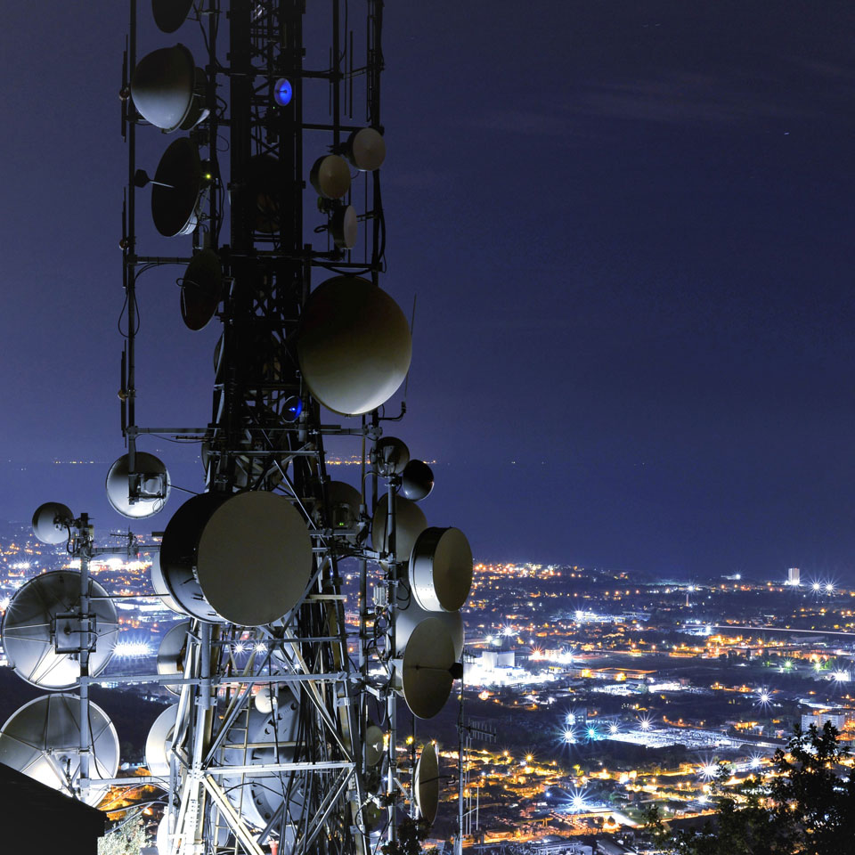 A telecommunications tower, antenna and satellite dish - a city's lights in the distance below