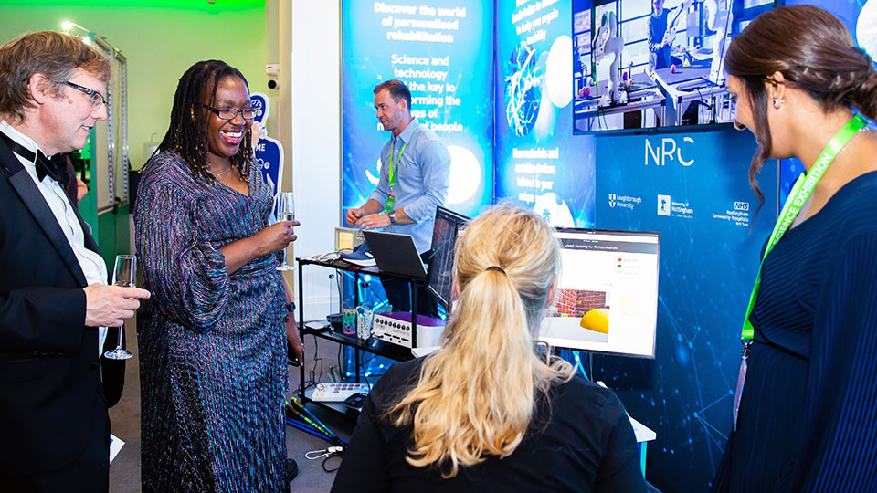 Two people holding drinks at the Royal Society Exhibition talking to two people, one sitting, while looking at a computer screen