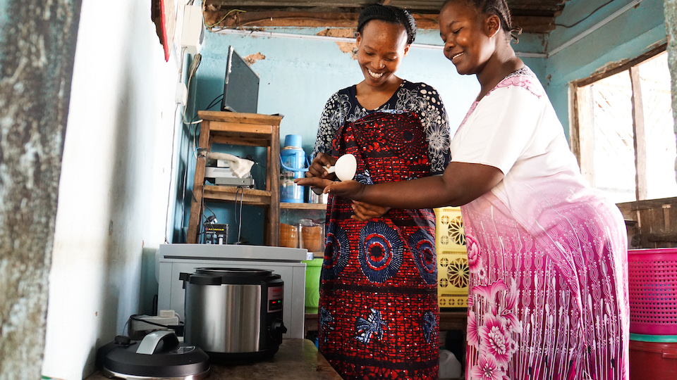 Two women standing in front of an electric cooking pot, they are measuring out ingredients using a large spoon.