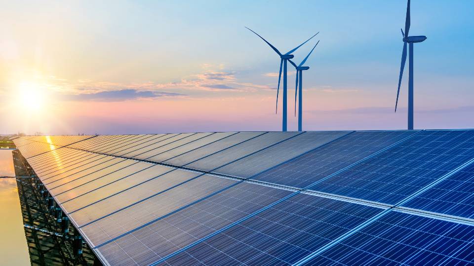 A row of solar panels with wind turbines in the background