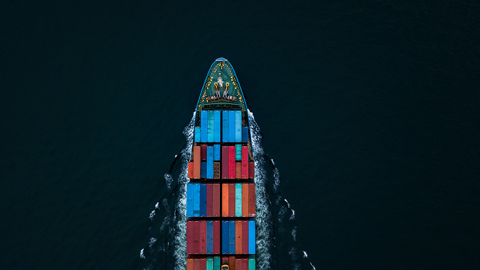 Aerial view of a container cargo ship travelling through dark ocean waters carrying multicoloured shipping containers, representing global supply chains and logistics networks.