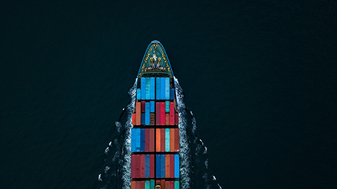 Aerial view of a container cargo ship travelling through dark ocean waters carrying multicoloured shipping containers, representing global supply chains and logistics networks.