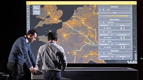Two people standing in front of a large interactive digital display showing a map-based supply chain or logistics network dashboard, with transport routes, data visualisations and operational controls visible on screen.